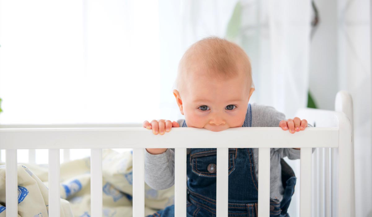 Babykamer-goed-inrichten Een baby knabbelt aan de leuning van een wit ledikantje in een felgekleurde Babyzimmer, gekleed in een grijs shirt en een spijkerbroek.