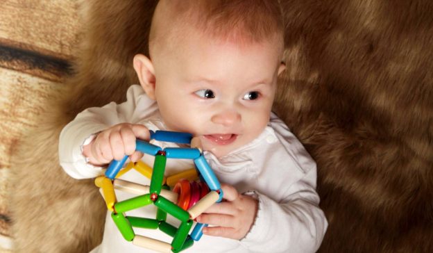 Tussen de 16 en 18 weken oud houdt een baby in een wit pakje een kleurrijk geometrisch speeltje vast terwijl hij op een harige ondergrond ligt.