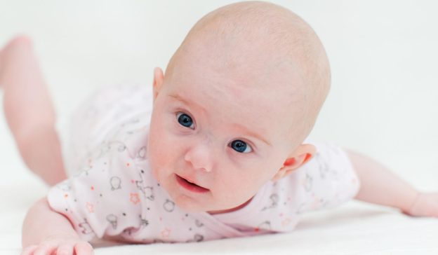 Een baby, ongeveer 6 tot 8 weken oud, in een wit pakje versierd met sterren- en uilenpatronen, ligt op zijn buik en kijkt nieuwsgierig voor zich uit.