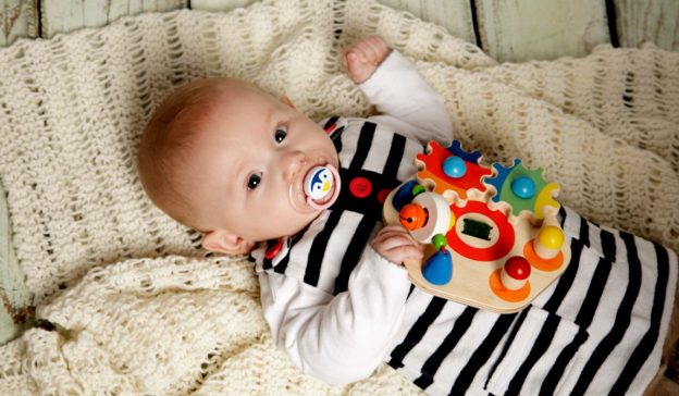 Een baby van 8 tot 12 weken oud ligt op een gebreid dekentje en houdt een kleurrijk speeltje vast, tevreden met een speen in zijn mond.