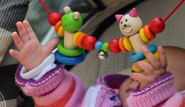 Baby reikt naar kleurrijk houten kikker- en kattenspeelgoed dat tussen de handen hangt, terwijl een autokinderwagen klaarstaat met netjes gerangschikte draagdoeken en draagzakken.