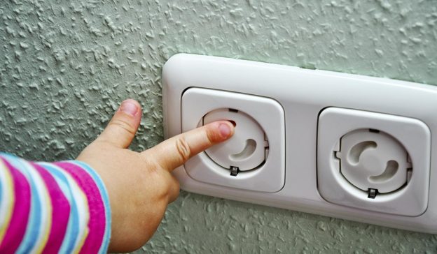 De hand van een kind, stevig in een kleurrijk gestreepte mouw, reikt naar een stopcontact dat beschermd is door veiligheidsklepjes: echt kindvriendelijk.