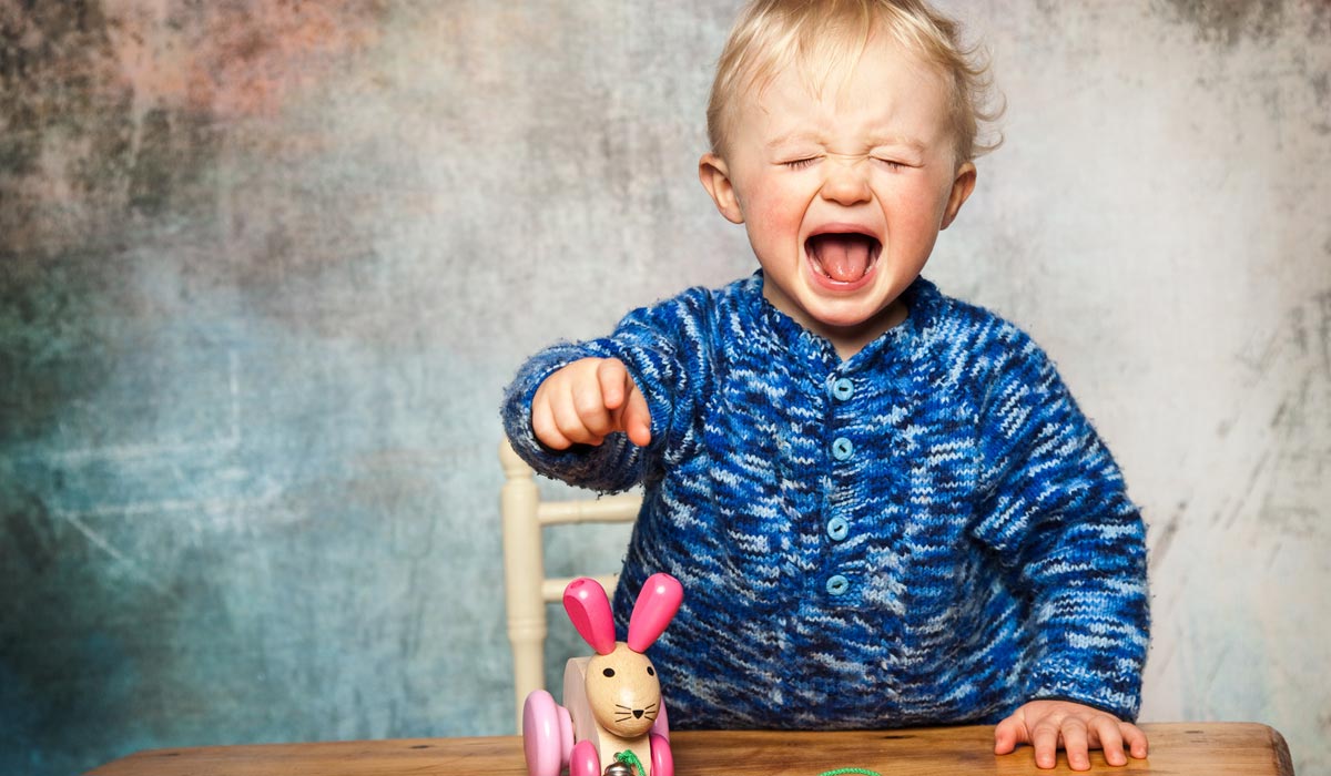 kind-heeft-driftbui Een lief kindje in een blauwe trui huilt en wijst ernstig naar een speelgoedkonijntje dat op tafel zit.