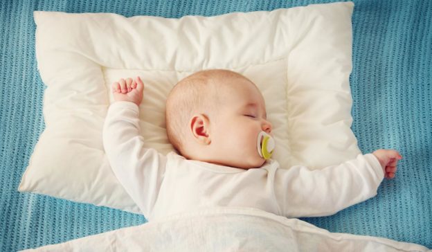 Een vredig slapende baby met een speen rust op een wit kussen en een blauwe deken, met de armen wijd gespreid, wat de perfecte slaapplek creëert voor zoete dromen.