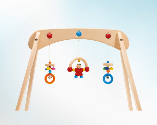 Stevige speeltrapeze van massief hout, met hangende kleurrijke ringen en een klein speelgoedfiguurtje, tegen een lichtblauwe achtergrond.