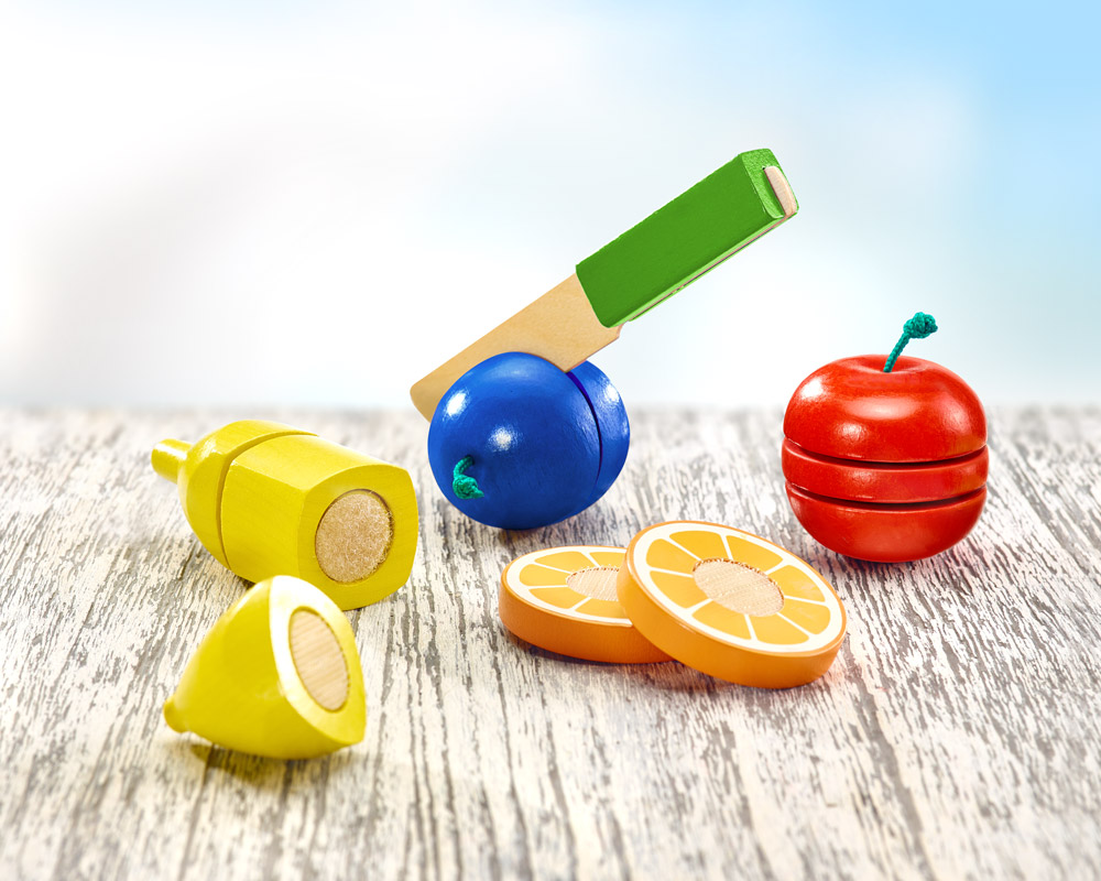 Houten speelgoedfruit, elk met gesneden fruit en klittenband voor eenvoudig snijden, en een messenlegger op een houten tafel tegen een blauwe achtergrond.