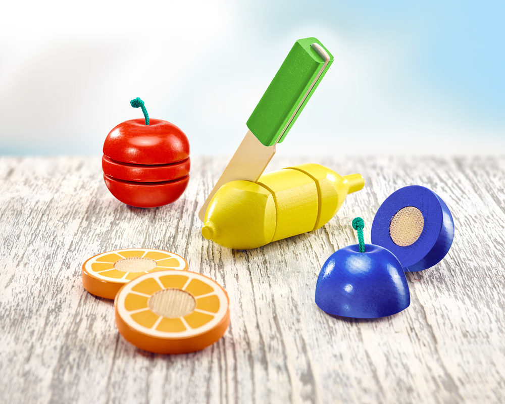 Houten speelgoedfruit en een mes rusten op een houten oppervlak, afgetekend tegen een helderblauwe lucht. Elk stuk is voorzien van gesneden fruit met klittenband voor interactief spel.
